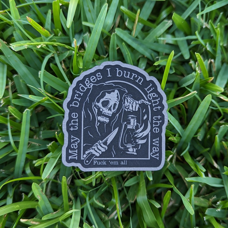 May include: A dark gray sticker featuring a grim reaper holding a lantern and a knife. The sticker has the text "May the bridges I burn light the way" and "Fuck 'em all". The sticker is placed on a bed of green grass.