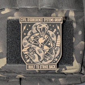May include: A black and tan tactical morale patch featuring a coiled rattlesnake wrapped around a globe. The text reads CIVIL DISOBEDIENCE SYSTEMS GROUP at the top and BUILT TO STRIKE BACK at the bottom. The design includes lightning bolts and stars.