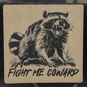 May include: A tan patch with a black and white illustration of a raccoon with its mouth open, showing its teeth. The raccoon is sitting on its haunches and has a fierce expression. The text "FIGHT ME COWARD" is printed below the raccoon.