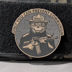 May include: A circular patch featuring a bear in a park ranger hat holding a rifle. The text "ONLY YOU CAN PREVENT COMMUNISM" surrounds the bear image. The patch has a tan border and a gray background.