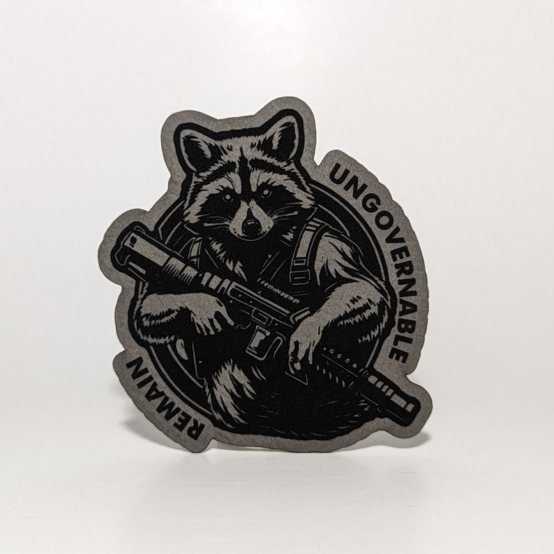 May include: A die-cut sticker featuring a raccoon holding a rifle. The raccoon is depicted in black and gray tones, with the words "UNGOVERNABLE" and "REMAIN" encircling the image. The sticker has a matte finish.