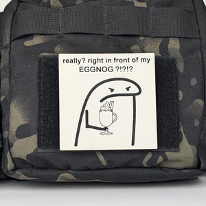 May include: A black and green camouflage fabric bag with a white patch that says "really? right in front of my EGGNOG ?!?" and a cartoon drawing of a person holding a glass of eggnog.