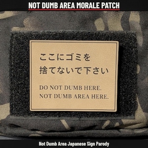 May include: A rectangular morale patch featuring a tan background with black text. The text reads in Japanese characters followed by the English phrases: DO NOT DUMB HERE. NOT DUMB AREA HERE. The patch is mounted on a black hook-and-loop fastener.
