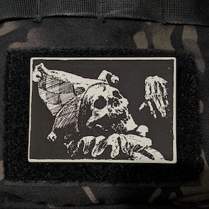 May include: A rectangular morale patch featuring a high-contrast black and white graphic illustration of a jester skull wearing a traditional multi-pointed cap with bells, accompanied by a skeletal hand gesture, all set against a black background.