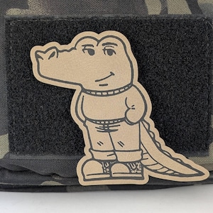 May include: A brown alligator cartoon character wearing a sweater and pants. The alligator has a long tail and is wearing shoes. The cartoon is on a black patch.
