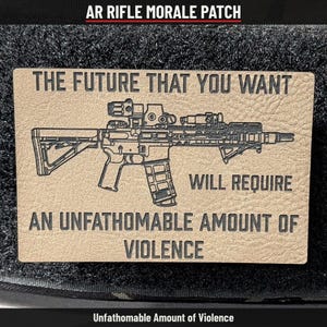May include: A rectangular tan morale patch featuring a black laser-engraved graphic of an AR-style rifle. The text reads: THE FUTURE THAT YOU WANT WILL REQUIRE AN UNFATHOMABLE AMOUNT OF VIOLENCE. The patch has a textured surface and black backing.