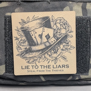 May include: A square patch with a top hat, playing cards, and floral elements. The text "LIE TO THE LIARS STEAL FROM THE THIEVES" is printed below the illustration. The patch is tan with a black and white design, and is set against a camouflage background.