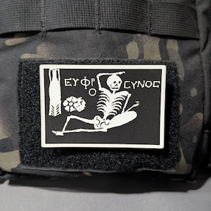 May include: A rectangular black and white morale patch featuring a white skeleton graphic in a reclining position. The design includes a floral element and the Greek text "ΕΥΦΡΟΣΥΝΟΣ" printed in a stylized font. The patch is attached to a dark camouflage fabric.
