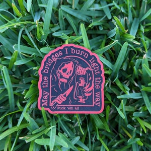 May include: A black sticker with a red border featuring a grim reaper holding a lantern and a knife. The text reads "May the bridges I burn light the way" and "Fuck 'em all". The sticker is on a bed of green grass.