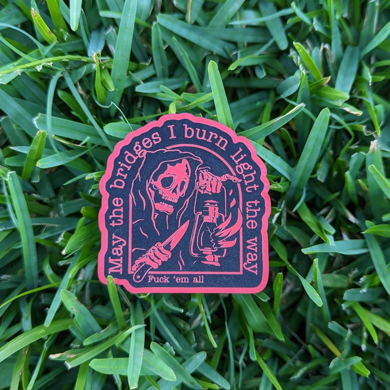 May include: A black sticker with a red border featuring a grim reaper holding a lantern and a knife. The text reads "May the bridges I burn light the way" and "Fuck 'em all". The sticker is on a bed of green grass.