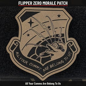 May include: A tan and black morale patch shaped like a shield. The design features a dolphin wearing a headset with lightning bolts radiating from it, stars, and the text "ALL YOUR COMMS ARE BELONG TO US" curved along the bottom edge.