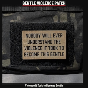 May include: A rectangular tan morale patch with black text that reads: NOBODY WILL EVER UNDERSTAND THE VIOLENCE IT TOOK TO BECOME THIS GENTLE. The patch is attached to a black hook-and-loop fastener on a dark camouflage patterned fabric background.