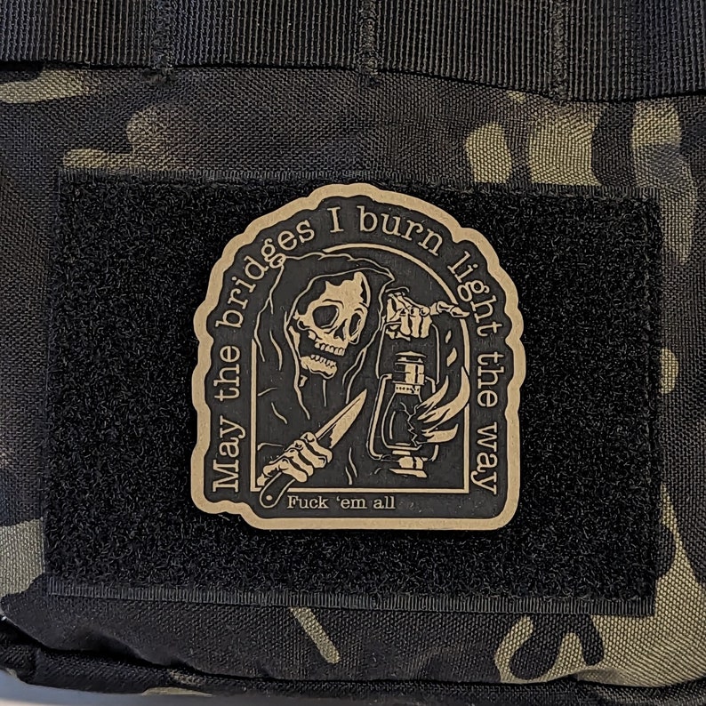 May include: A rectangular patch with a Grim Reaper design, holding a lantern and a knife. The patch has the text "May the bridges I burn light the way" and "Fuck 'em all". The patch is on a black velcro surface.