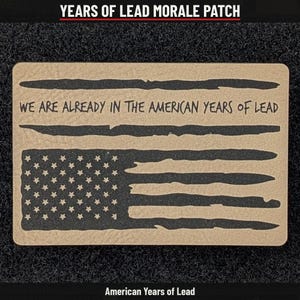 May include: A rectangular morale patch in tan and black featuring a stylized American flag design. The text WE ARE ALREADY IN THE AMERICAN YEARS OF LEAD is printed across the center. The flag includes a field of stars and horizontal stripes.
