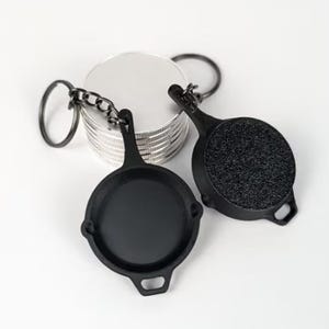 May include: Two black frying pan keychain charms with a silver chain and ring. The frying pans have a textured surface.
