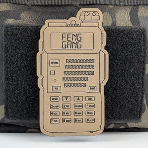 May include: A brown and black two-way radio sticker with the text "FENG GANG" on the display. The sticker has a variety of buttons and labels, including "VFO/MR", "A/B", "BAND", "MENU", "STEP", "TXP", "SAVE", "SCAN", "VOX", "WW", "ABR", "SOL", "TDR", "BEEP", and "TOT".