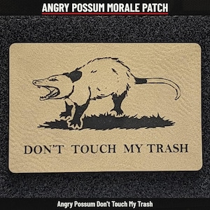 May include: A rectangular morale patch with a tan background and a black graphic of an angry, hissing opossum standing on grass. The text DON'T TOUCH MY TRASH is printed in black capital letters below the opossum illustration.