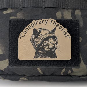 May include: A patch with the text "Conspiracy Theorist" and a black and white illustration of a cat wearing a hat. The patch is attached to a black fabric square, which is on a camouflage background.