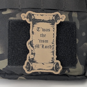May include: A brown scroll patch with the text "T'was the 'tism M'Lord" in black lettering. The patch is attached to a black and green camouflage fabric.