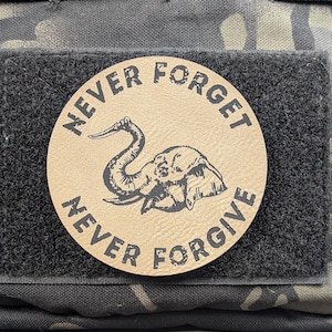 May include: A round, tan patch with the words "NEVER FORGET" and "NEVER FORGIVE" in black, surrounding a black elephant illustration. The patch is attached to a black velcro square, set against a camouflage background.