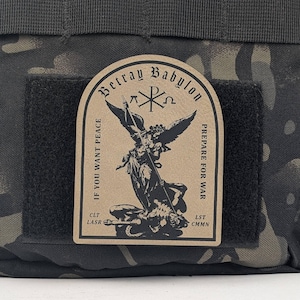 May include: A brown patch with the text "Betray Babylon" and "Prepare for War" on it. The patch features a black and white illustration of an angel defeating a demon. The text "If you want peace" is on the left side of the patch and "CLT LASR" and "LST CMNN" are on the bottom.