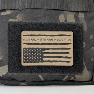 May include: A tan-colored patch with a black and white American flag and the text "WE ARE ALREADY IN THE AMERICAN YEARS OF LEAD" on a black and green camouflage background.
