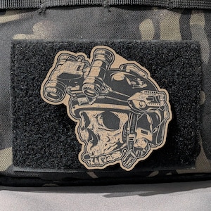 May include: A tan and black patch depicting a skull wearing a tactical helmet and night vision goggles. The patch is attached to a black velcro surface, with a camouflage pattern in the background.