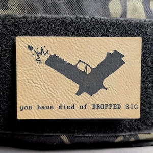 May include: A tan leather patch with a black silhouette of a handgun firing a bullet. The text "you have died of DROPPED SIG" is printed below the image.