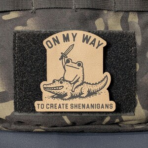 May include: A tan rectangular morale patch with black text and a black line art illustration. The text reads ON MY WAY TO CREATE SHENANIGANS. The illustration depicts a frog holding a sword while riding on the back of a large alligator.