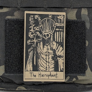 May include: A brown card with a black and white illustration of a skeletal figure wearing a robe and a crown. The figure is holding a cross and is standing in front of a building. The text "The Hierophant." is printed at the bottom of the card.