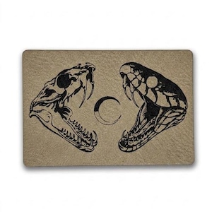 May include: A rectangular, tan-colored doormat featuring a black illustration of two snake heads facing each other, with a crescent moon in the center. The snake heads have open mouths and visible fangs.