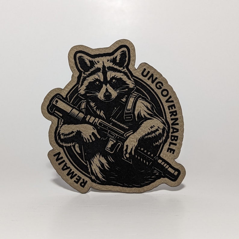 May include: A die-cut sticker featuring a raccoon holding a rifle. The raccoon is black and the sticker has a tan background. The words "UNGOVERNABLE" and "REMAIN" are printed on the sticker.