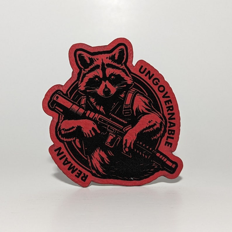 May include: A red and black sticker featuring a raccoon holding a rifle. The raccoon is inside a black circle with the words "UNGOVERNABLE" and "REMAIN" in a red font. The sticker has a die-cut edge.