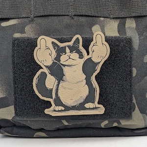 May include: A black and white cat with a grumpy expression is giving the middle finger with both hands. The cat is on a brown patch with a black border. The patch is on a camouflage background.