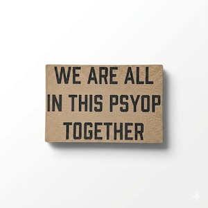 May include: A rectangular, tan-colored sign with the phrase "WE ARE ALL IN THIS PSYOP TOGETHER" printed in bold, black capital letters. The sign has a textured surface and is set against a white background.