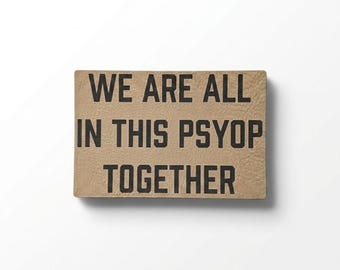 May include: A rectangular, tan-colored sign with the phrase "WE ARE ALL IN THIS PSYOP TOGETHER" printed in bold, black capital letters. The sign has a textured surface and is set against a white background.