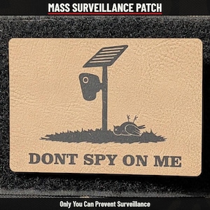 May include: A rectangular tan patch featuring a black graphic of a security camera on a pole with a solar panel and a dead bird on the ground. The text reads MASS SURVEILLANCE PATCH, DONT SPY ON ME, and Only You Can Prevent Surveillance.