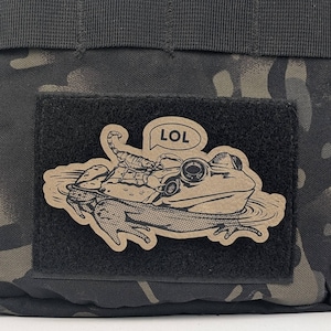 May include: A brown patch with a black border featuring a cartoon frog with a scorpion on its back. The frog is wearing goggles and has a speech bubble above its head that says "LOL".