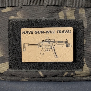 May include: A rectangular tan morale patch featuring a black line drawing of a submachine gun and the text HAVE GUN-WILL TRAVEL in bold black capital letters. The patch is attached to a black hook-and-loop fastener on a camouflage fabric.