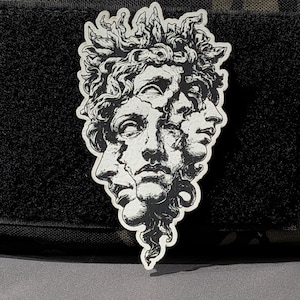May include: A black and white sticker featuring a classical sculpture of a face with multiple overlapping profiles. The sticker has a detailed, artistic design with a textured appearance, suitable for various applications.
