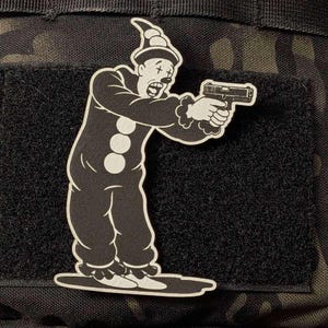 May include: A black and white morale patch featuring a cartoon clown wearing a pointed hat and a ruffled suit, holding a handgun with both hands. The clown has a sad expression with cross-shaped markings over the eyes and three large buttons.