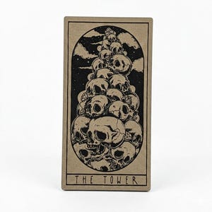 May include: A tarot card featuring a black and white illustration of a tower of skulls against a cloudy sky. The card is rectangular with the words "THE TOWER" printed at the bottom.