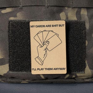 May include: A rectangular tan morale patch with a black hook-and-loop backing. The patch features a black line drawing of a hand holding a fan of playing cards, with the text MY CARDS ARE SHIT BUT I'LL PLAY THEM ANYWAY printed in black.