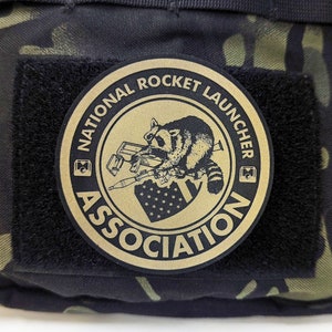 May include: A tan round patch with black text and a black and white illustration of a raccoon holding a rocket launcher. The text reads "National Rocket Launcher Association".