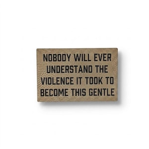 May include: A rectangular wooden sign with the text "NOBODY WILL EVER UNDERSTAND THE VIOLENCE IT TOOK TO BECOME THIS GENTLE" in black lettering. The sign has a light brown, textured background.