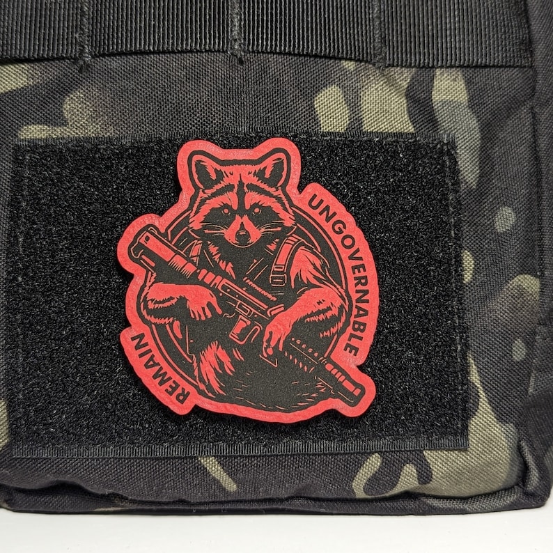May include: A red and black patch featuring a raccoon holding a rifle. The patch is affixed to a black fabric with a hook and loop fastener. The words "REMAIN" and "UNGOVERNABLE" are printed on the patch.