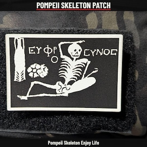 May include: A rectangular black and white patch featuring a skeleton graphic in a reclining position. The design includes Greek lettering and a floral element. The text POMPEII SKELETON PATCH appears above, and Pompeii Skeleton Enjoy Life appears below.