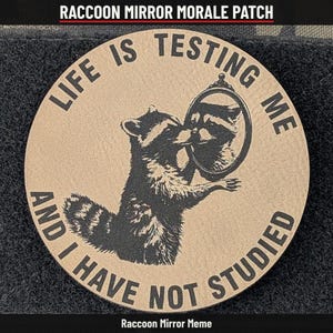 May include: A circular tan morale patch featuring a black graphic of a raccoon looking into an oval mirror. The text surrounding the image reads, "LIFE IS TESTING ME AND I HAVE NOT STUDIED." The patch has a textured surface and black border.