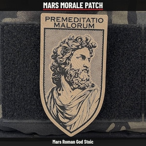 May include: A tan morale patch featuring a black line art illustration of a bearded man with curly hair in a toga. The text PREMEDITATIO MALORUM is printed in black at the top. The shield-shaped patch is attached to a black fabric surface.