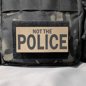 May include: A tan rectangular patch with the words "NOT THE POLICE" in black block letters is affixed to a black tactical bag with a camouflage pattern. The patch is attached with a black hook and loop fastener.